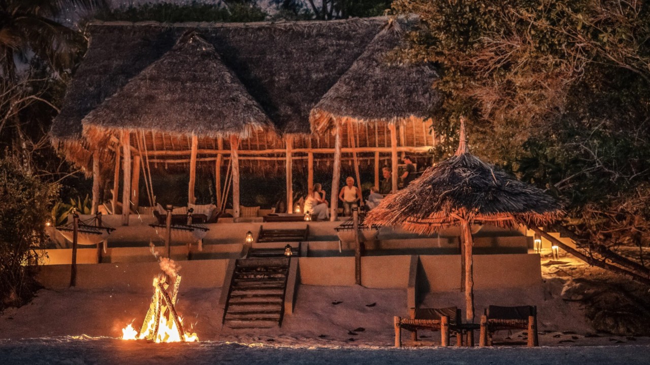 Nighttime at The Beach Bar at The Manta Resort, with a cozy fire burning on the sand and ambient lighting under the thatched roof.