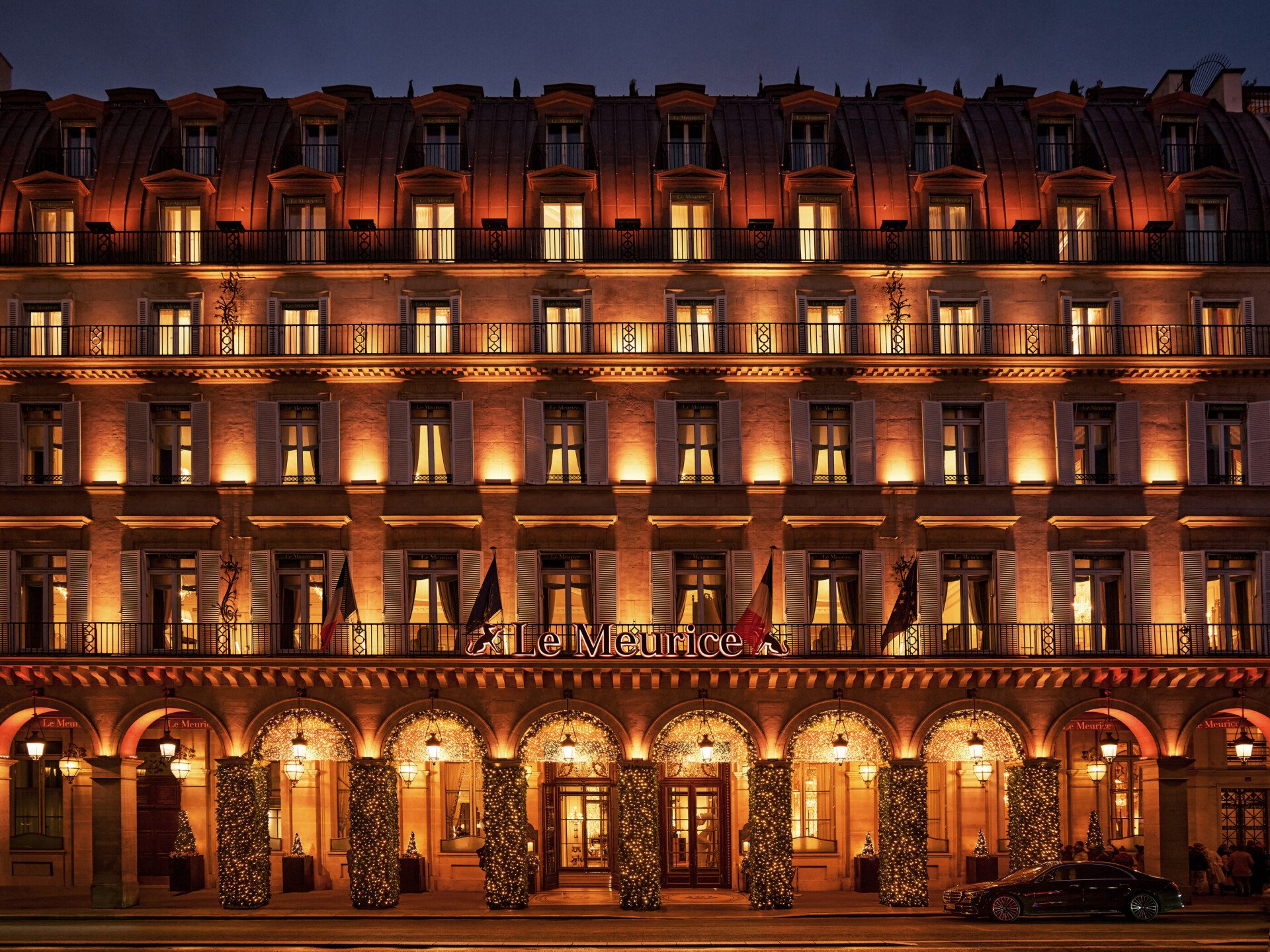 Le Meurice Paris hotel's illuminated facade on Rue de Rivoli, decorated with festive lights and holiday wreaths for the Christmas season.