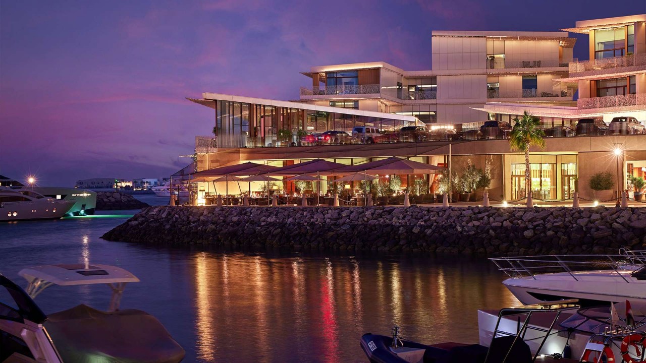 Bvlgari Resort Dubai is seen at sunset from Jumeira Bay, showcasing its Mediterranean architecture and luxury yachts in the marina against a vibrant sky.