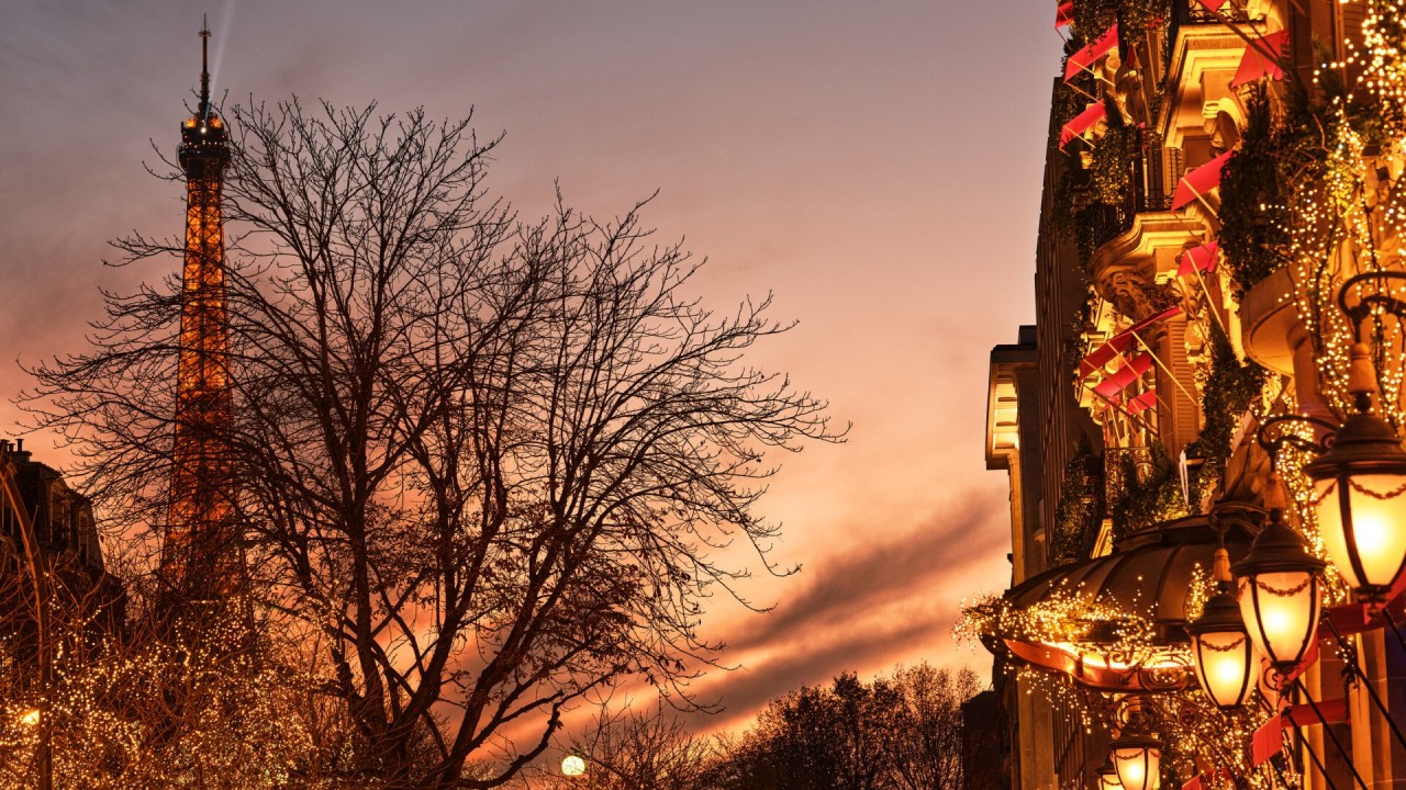 The historic facade of Hôtel Plaza Athénée, decorated for Christmas at sunset with the Eiffel Tower in the background.