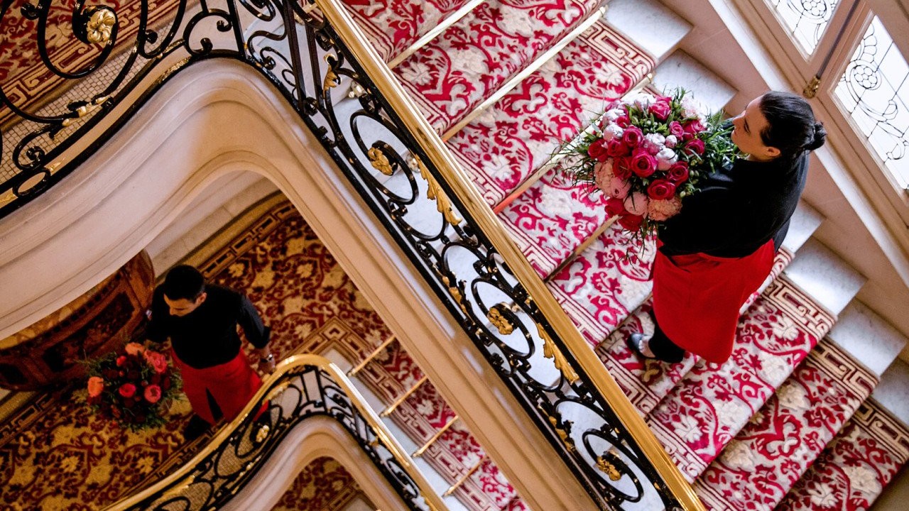 Staff members carrying vibrant red and pink floral bouquets through the legendary red and gold staircase at Hôtel Plaza Athénée, Paris.