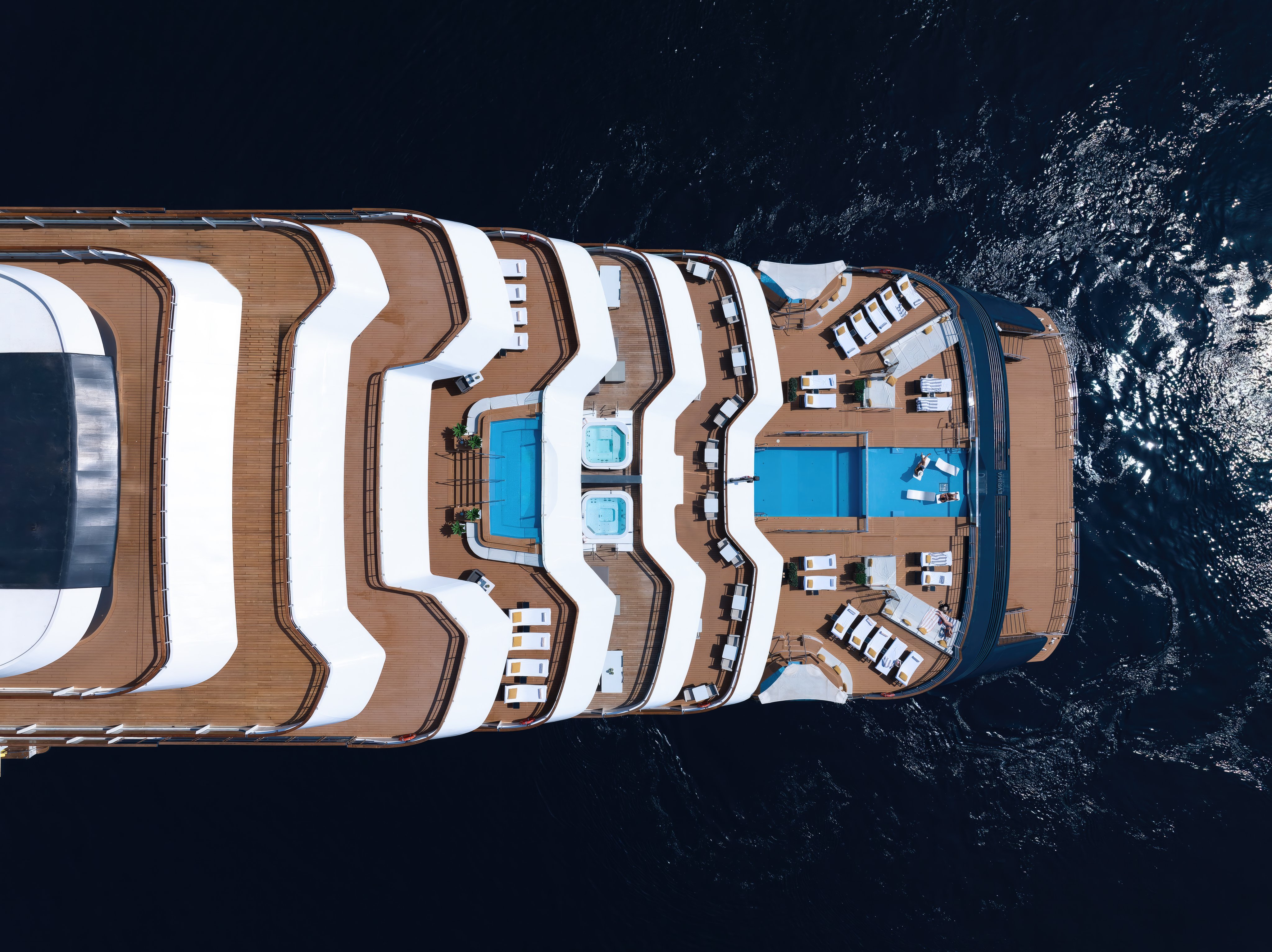 An aerial view of the Evrima yacht, showcasing its sleek design and expansive sun decks against a stunning coastline.