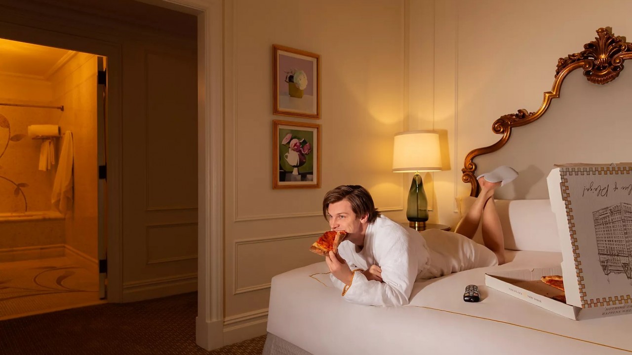 A guest at The Plaza Hotel relaxes in a plush robe on the bed, enjoying a slice of pizza while watching Home Alone, recreating the iconic movie night scene.