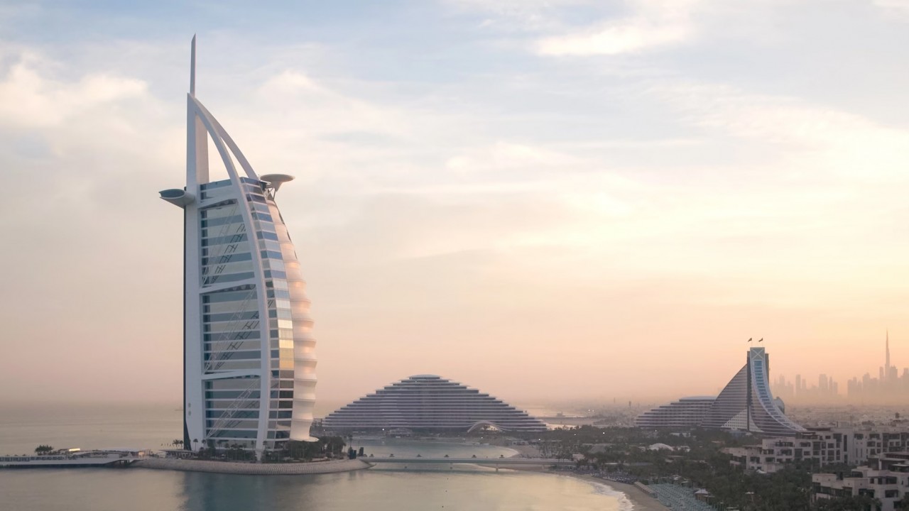 Aerial view of the iconic Burj Al Arab Jumeirah sail-shaped hotel on its private island, with the Dubai Marina skyline and coastline visible in the background.